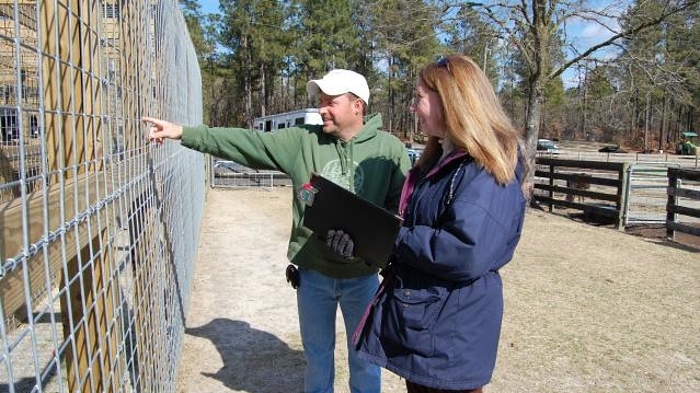 Female veterinarian holding a clipboard. She's standing next to a man pointing towards a fenced enclosure.