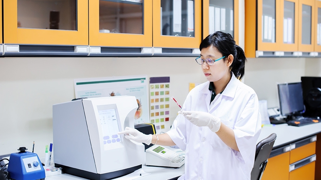 A woman wearing a lab coat looks at a test strip and presses a button on a nearby computer