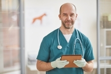 A man with green scrubs, a clipboard, and medical gloves holds a clipboard and looks at the camera.